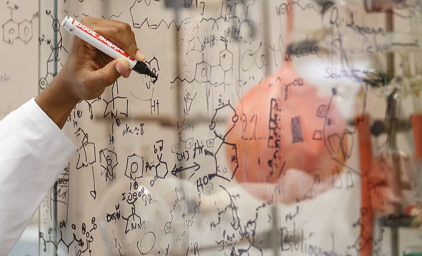 Hand eines Forschers, der chemische Formeln auf Glaswand notiert
